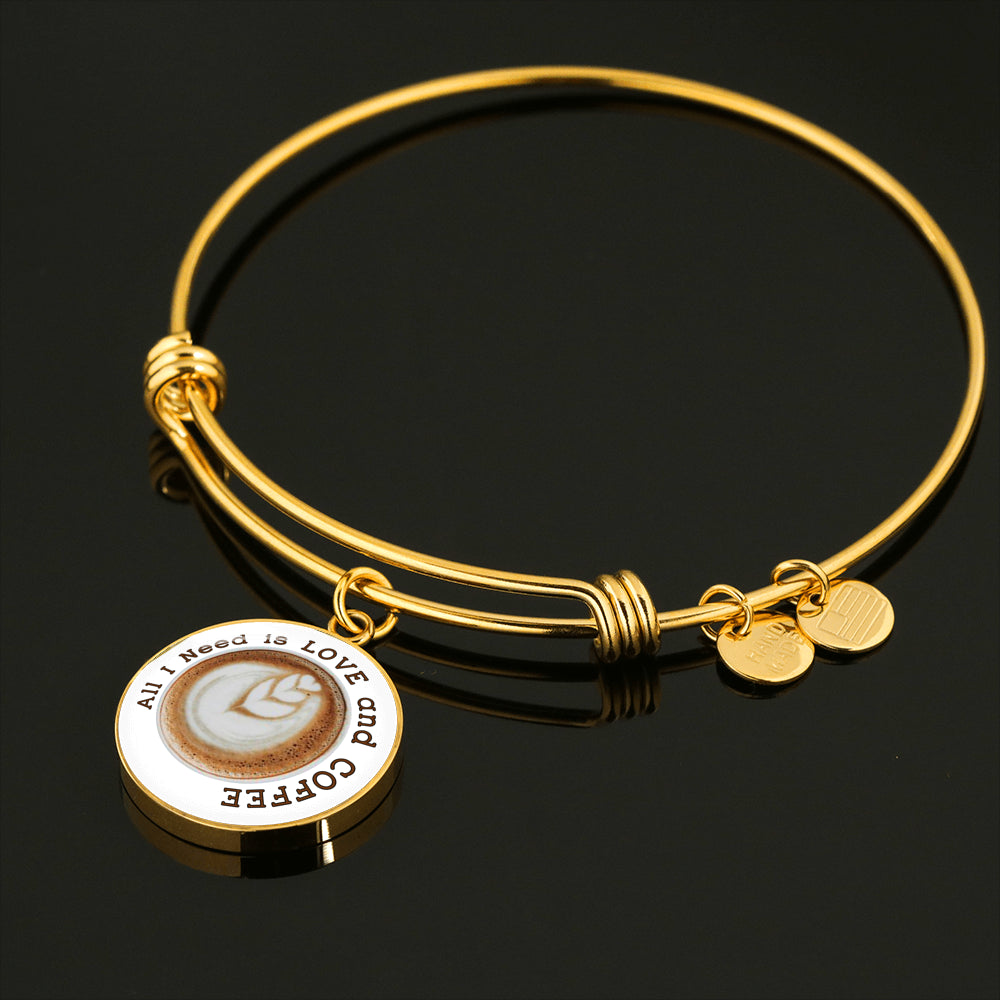 Gold Bangle lies on black, attached Love Coffee Charm says "All I Need is Love and Coffee" around a photo of coffee art depicting 3 hearts coming out of the coffee froth.