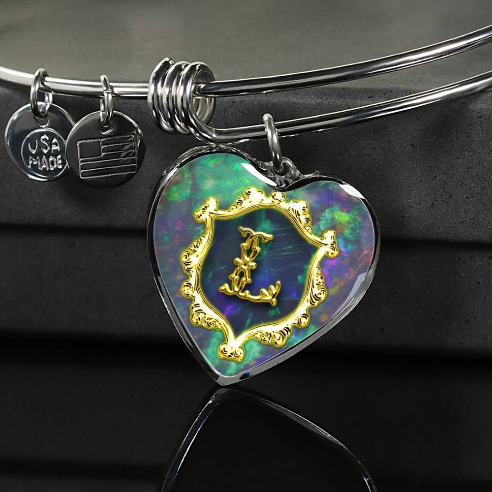close up of Heart Pendant Monograms L Alphabet Initial Polished stainless steel wire bangle; pendant is heart shaped with antique letter L surrounded by a shield, both appear to depict a gold-like finish, over a photo of green blue opal. draped over a black box